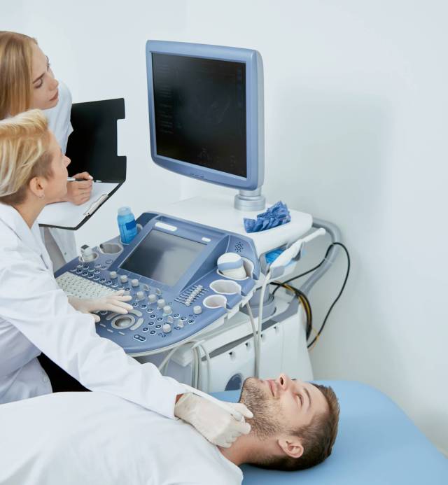 Side view of three people on medical consultation. Pofessional doctor sitting working examining patient looking at monitor and young assistant standing near and helping her.