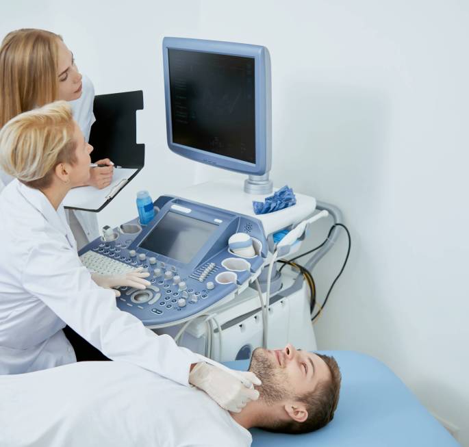 Doctor and assistant poviding medical examination. Side view of three people on medical consultation. Pofessional doctor sitting working examining patient looking at monitor and young assistant standing near and helping her.
